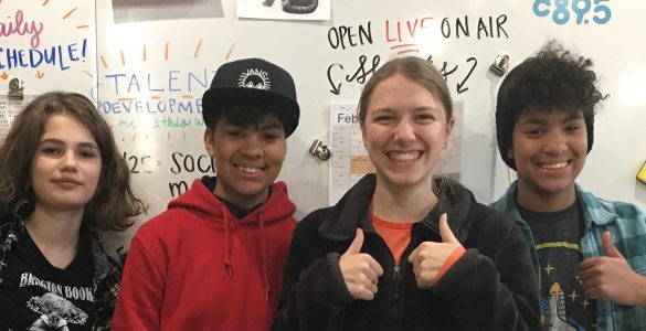 Four students standing in front of a large classroom white board. Two students are dark-skinned and appear to be identical twins. The other two students have lighter skin. Two students have wide smiles and are giving the thumbs up sign. Words on the white board behind the students include: daily schedule, talent development, and live on-air.