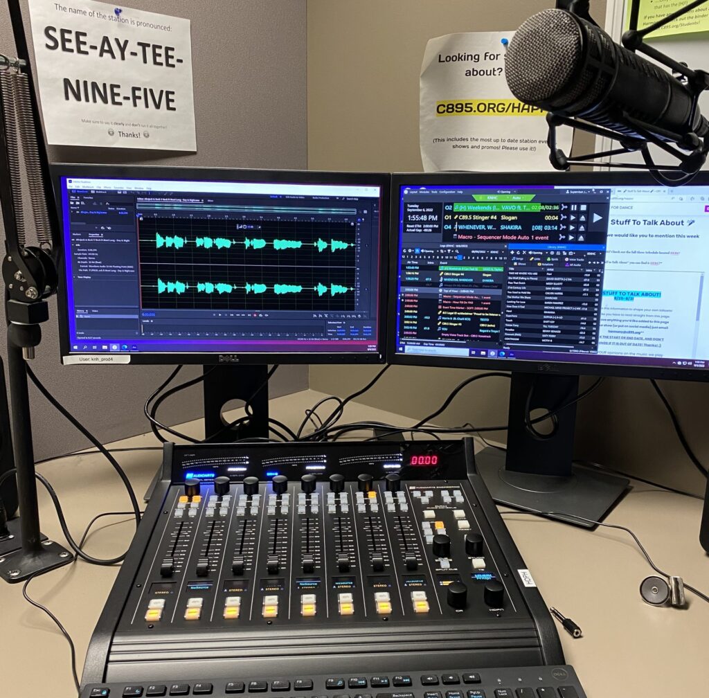 A picture of a broadcast audio mixing board with two computer monitors behind it and a sign which spells out how to say C89.5 aloud.