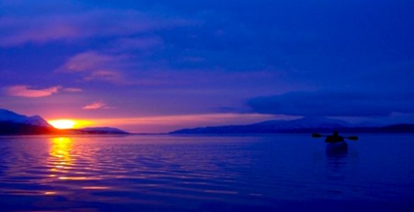 A person kayaking on a lake at sunset, with mountains in the background. The sky is a dark blue.