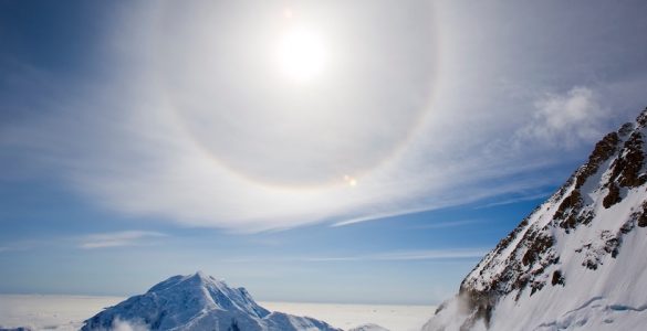 A halo around the sun, over a snowy landscape, with mountain peaks in the foreground. The sky is party cloudy but bright.