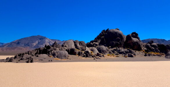 In the foreground is a flat dry lake bed, with a outcropping of rocks that somewhat resemble a grandstand at a race track. The sky is blue and there are mountains further in the desert background.