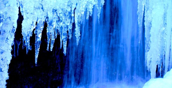 Icicles in the foreground with a waterfall in the background. The ice and water have a blue tint to them.