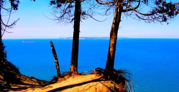 And sand-covered cliff edge with deciduous trees. The cliff is overlooking a large lake that is deep blue. The sky in the background is blue. There is some haze in the distance.