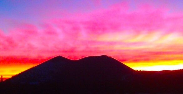 The outline of a backlit mountain. In the background is a colorful sky, illuminated by bright pink, yellow and purple clouds at sunset.