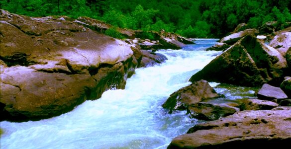 A river with water moving rapidly through a rocky landscape, creating white rapids. Other than the rapids, the water is blue. The rocks are brown and shiny from the mist of the water. In the background is a hillside with green trees. The sky is clear and blue.