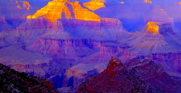 View of a canyon extending to the horizon. The canyon is shaded with sunlight reaching the peaks of the mountains within the canyon. In the foreground is the shaded edge of a cliff. The sky is blue with some white clouds on the horizon.