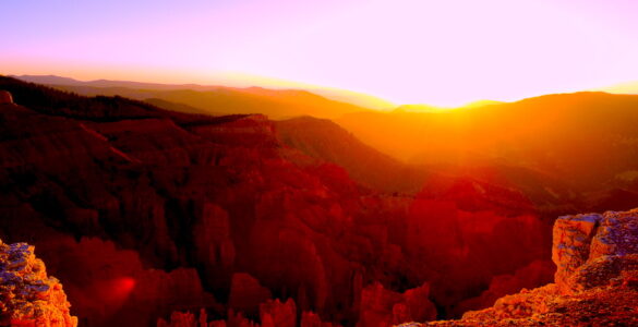 The sun is setting behind a ridge of mountains. In the foreground is the edge of a cliff exposing the jagged, but beautiful landscape below. Another range of mountains falls between the cliff and the ridge with the sunset. The landscape is orange, and the sky is shades of green and purple, with a yellow and orange sunset.