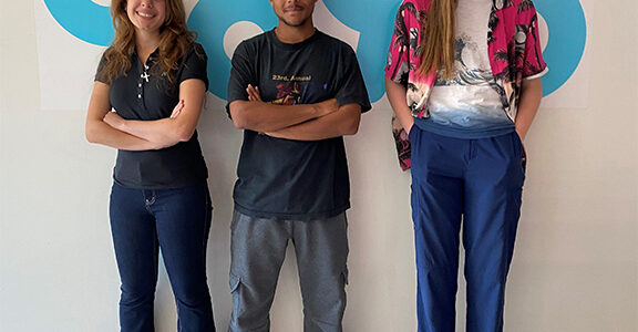 A photo of three teenagers standing in front of a white wall with the C89.5 logo. The person on the left has long brown hair and tan skin and has their arms folded. The person in the middle has dark skin and black curly hair and their arms crossed. The person on the right has long brown hair, glasses, pale skin and has their hands in their pockets. They all looked relaxed and are smiling.