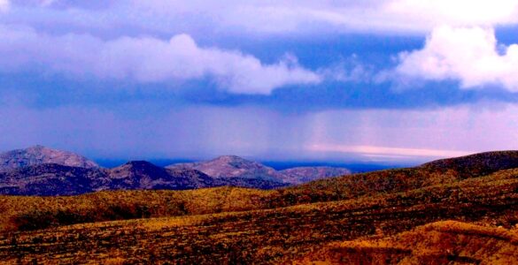 A desert landscape featuring a distance rainstorm. In the foreground are short, orange hills without vegetation. Behind that are taller hills that appear blue, and are also without vegetation. Behind that is a blue cloudy sky, with two curtains of rain reaching the landscape.