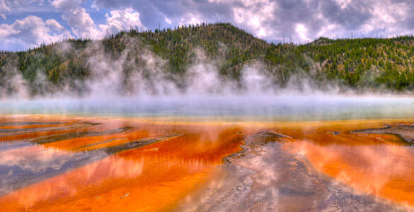 A volcanically active body of water with steam rising above. The water in the foreground appears orange or rust colored. The water further away appears to be blue. In the background are hills with green and brown foliage. The sky conditions are mostly cloudy, with sections of blue peeking through.