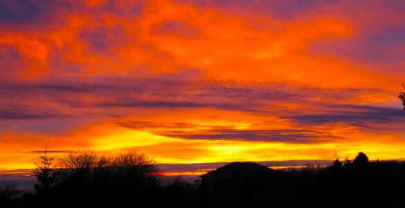 A bright sunset is illuminated on a mostly cloudy sky, with hues of orange and yellow. Parts of the clouds are dark blue. In the foreground, on the lower-third, is a building and some deciduous trees with no leaves, and a sole evergreen tree. There is also a cabin the the foreground. The foreground is shown as mostly an outline, as the sun is not providing light on that area.