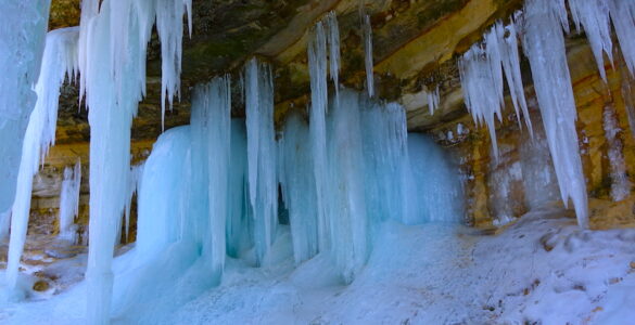 Icicles hanging from below an escarpment. The icycles are robust and light blue. There is white ice on the ground.