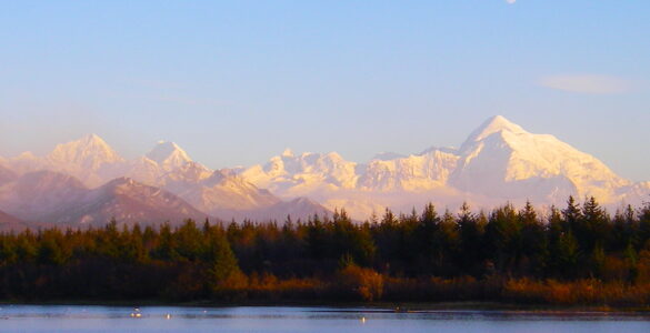 In the foreground is a lake with calm blue waters. The shore of the lake is forested, mostly with evergreen trees. In the background are tall mountains, the ones the furthest away are snow covered. The sky is blue with a three-quarter moon showing in the daylight.