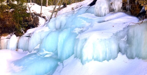 A fully frozen cascading waterfall covered mostly in snow. The waterway ice is blue. Trees and bushes surround the waterway.