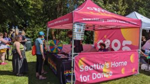 A group of people is gathered at an outdoor community event, interacting near booths with colorful banners, including one labeled "c895," under a clear blue sky.