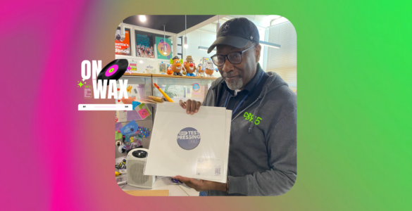 A person in a cap and sweater holds a vinyl record album labeled "Test Pressing" in a vibrant, colorful store filled with vinyl records and music paraphernalia, with a neon sign saying "ON WAX" visible in the background.