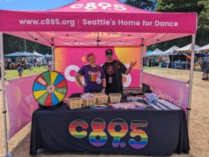 Two individuals standing behind a promotional booth for C89.5, labeled as "Seattle’s Home for Dance," at an outdoor event. The booth features a bright pink banner, a spinning prize wheel, and various promotional items on display, including brochures and branded merchandise.