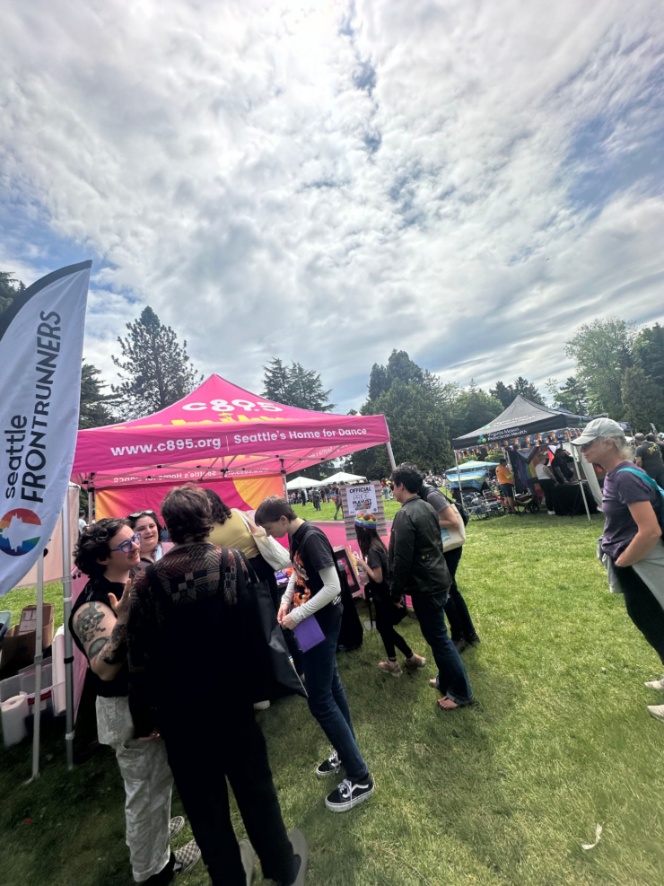 An outdoor event with several garden tents and promotional banners. To the left is a blue banner reading "Seattle Runners," and centrally is a vibrant pink tent with a banner "C89.5 - Seattle's Home for Dance," where people are gathered around. The location is a park-like setting with lush green grass and trees. The sky is partly cloudy. People of various ages are standing and engaging with one another, some examining items on the tables under the tents.