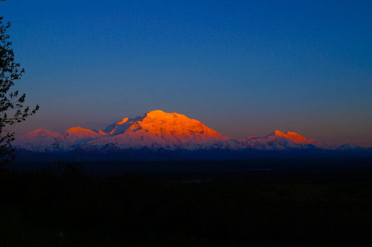 A distant snow-capped peak under the diffused reddish sunlight, with soft atmospheric shadows, contrasting against a dark nighttime foreground.