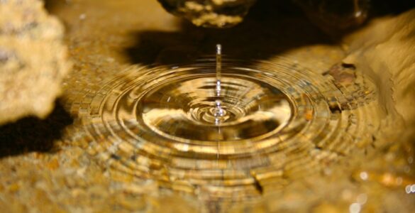 A clear pool of water inside a cave. A single drop disrupts the still surface, creating ripples. The bottom of the pool is covered in sediment and surrounded by brownish cave walls.