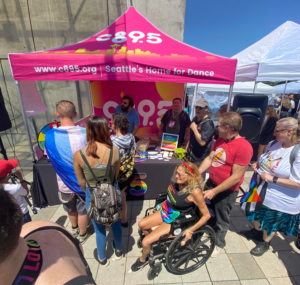 A vibrant promotional booth with the text "C89.5 - c895.org Seattle's Home for Dance" on a pink banner. Several people are gathered around the booth, with one person in a wheelchair engaging in conversation with an individual behind the booth. The booth is decorated with a rainbow-colored panel, and various items like stickers and brochures are on display. The background shows a clear blue sky and other festival booths.