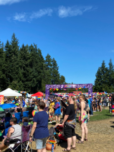 Outdoor scene at a community festival, with numerous people gathered on a sunny day. The crowd varies in age and attire, focused around a vibrant booth with a sign labeled "Pride" in purple letters. Trees line the background under a clear blue sky. Various colorful flags and casual outdoor chairs populate the area, indicating a festive atmosphere.