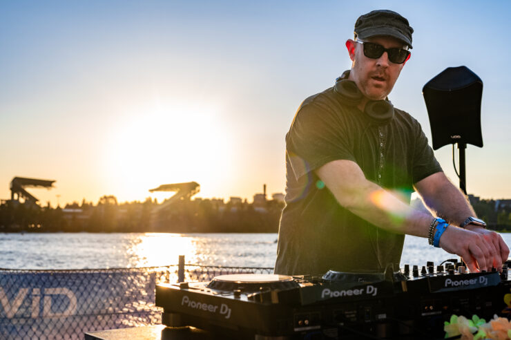 A DJ at CD-Js wearing sunglasses and looking down with a sunset behind them on the water