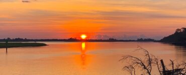 A sunrise casts an orange glow over a cloudy sky and a tranquil lake, with trees lining the shore.