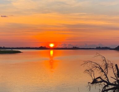 A sunrise casts an orange glow over a cloudy sky and a tranquil lake, with trees lining the shore.