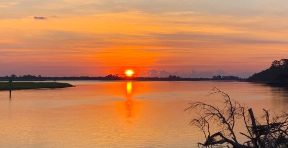 A sunrise casts an orange glow over a cloudy sky and a tranquil lake, with trees lining the shore.