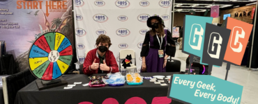 Two individuals at a CB95 promotional booth at a convention, featuring a prize wheel, merchandise, and banners. The booth is adorned with a 'Start Here' sign and the motto 'Every Geek, Every Body'.