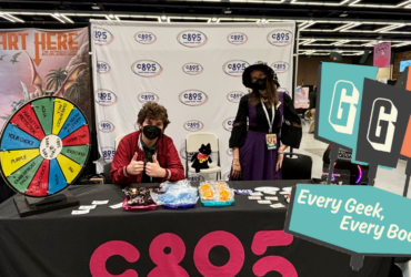 Two individuals at a CB95 promotional booth at a convention, featuring a prize wheel, merchandise, and banners. The booth is adorned with a 'Start Here' sign and the motto 'Every Geek, Every Body'.