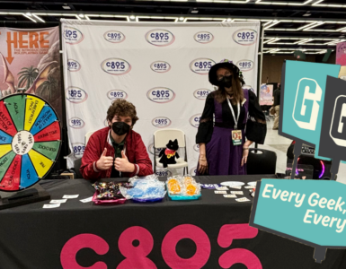 Two individuals at a CB95 promotional booth at a convention, featuring a prize wheel, merchandise, and banners. The booth is adorned with a 'Start Here' sign and the motto 'Every Geek, Every Body'.