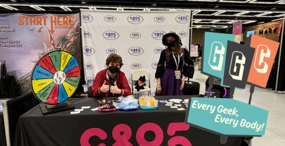 Two individuals at a CB95 promotional booth at a convention, featuring a prize wheel, merchandise, and banners. The booth is adorned with a 'Start Here' sign and the motto 'Every Geek, Every Body'.