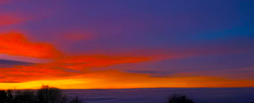 A view of a valley with clouds covering the valley floor, shortly after sunset. The sky is blue with shades of orange and red.