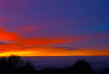 A view of a valley with clouds covering the valley floor, shortly after sunset. The sky is blue with shades of orange and red.