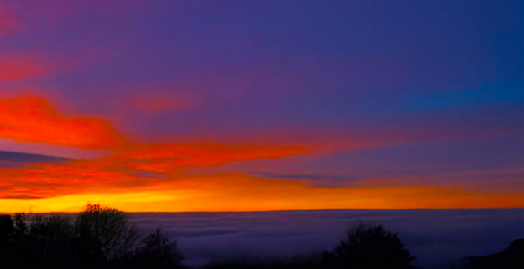 A view of a valley with clouds covering the valley floor, shortly after sunset. The sky is blue with shades of orange and red.