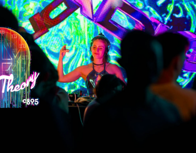 A DJ performs at a nightclub, surrounded by colorful lighting and an engaged crowd, enhancing the lively atmosphere.