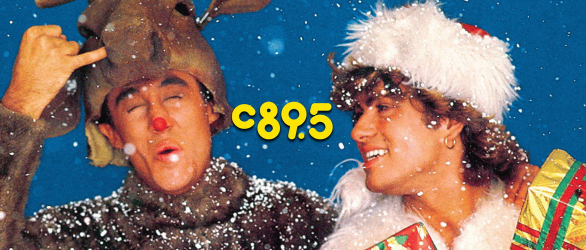 Promotional image from the song 'Last Christmas' by Wham! featuring a person in a reindeer costume winking and making a peace sign, and another person in a white fur hat smiling while holding a stack of gifts, both surrounded by falling snow.