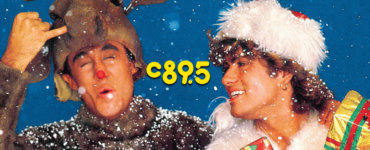 Promotional image from the song 'Last Christmas' by Wham! featuring a person in a reindeer costume winking and making a peace sign, and another person in a white fur hat smiling while holding a stack of gifts, both surrounded by falling snow.