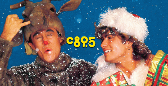 Promotional image from the song 'Last Christmas' by Wham! featuring a person in a reindeer costume winking and making a peace sign, and another person in a white fur hat smiling while holding a stack of gifts, both surrounded by falling snow.