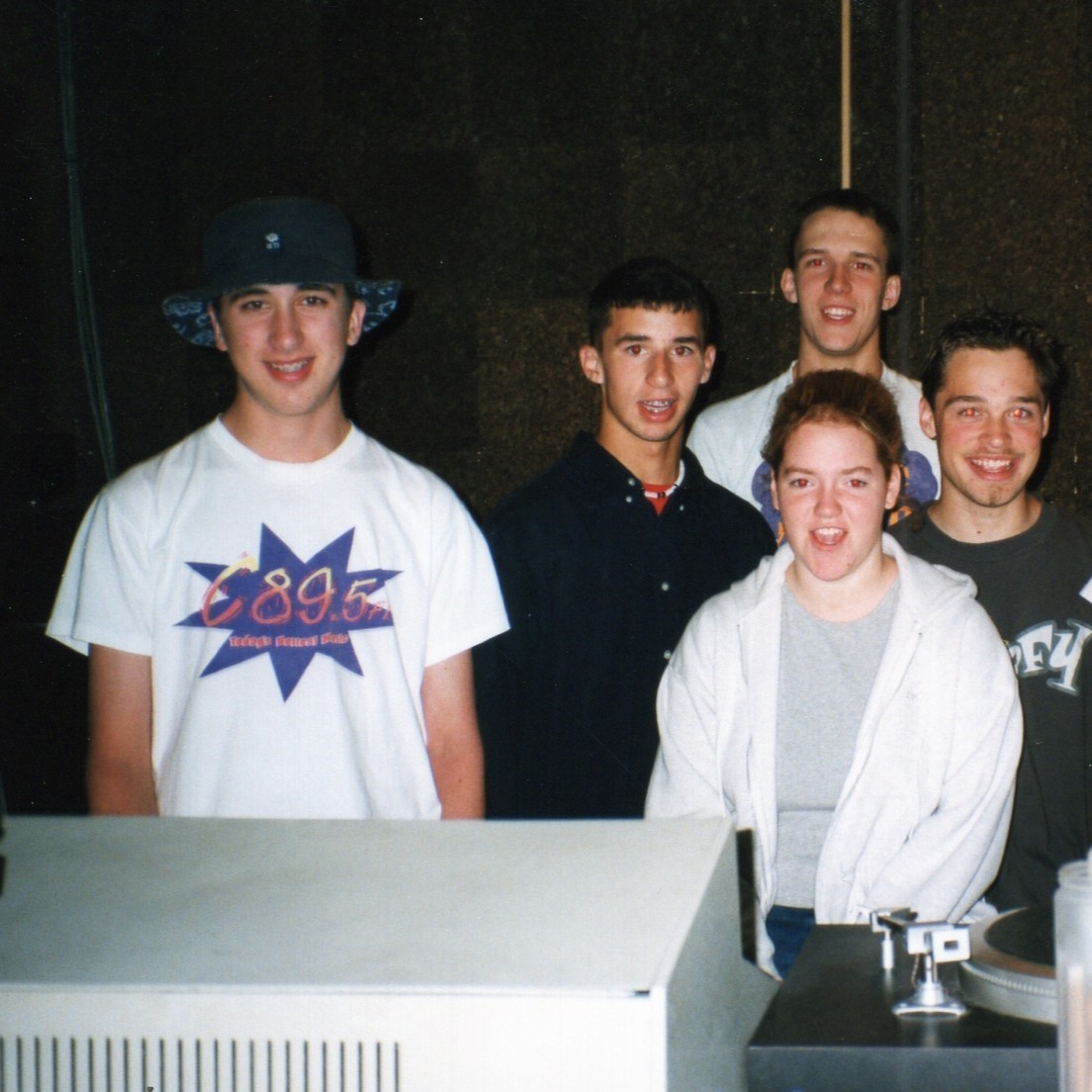 Five individuals smiling at a computer station, one wearing a hat and a t-shirt marked with "895 FM"