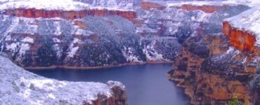 Snow-covered cliffs of a deep canyon with a lake on the canyon floor, under a cloudy sky