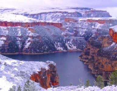 Snow-covered cliffs of a deep canyon with a lake on the canyon floor, under a cloudy sky