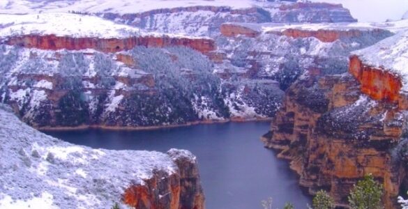 Snow-covered cliffs of a deep canyon with a lake on the canyon floor, under a cloudy sky