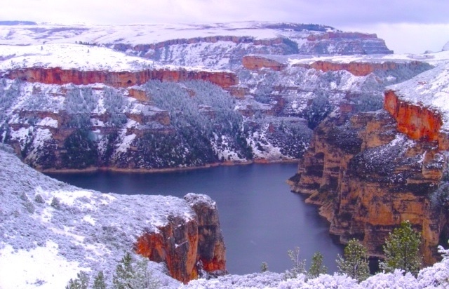 Snow-covered cliffs of a deep canyon with a lake on the canyon floor, under a cloudy sky