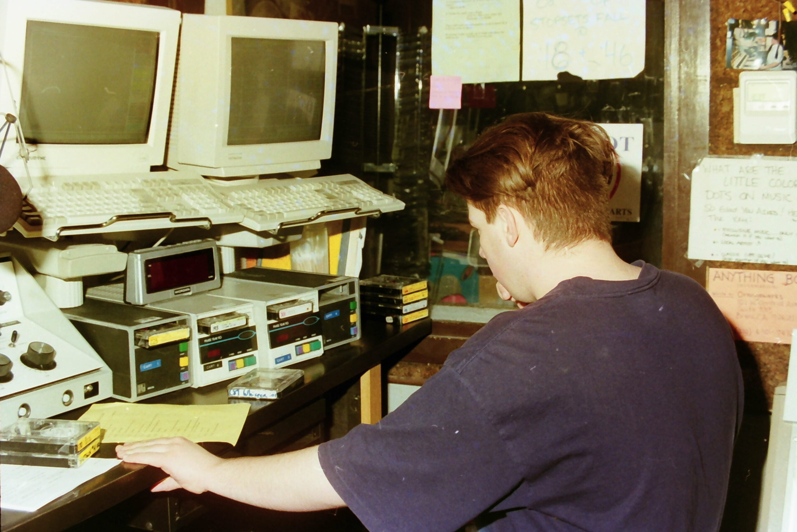 Person reading a document in a radio studio with vintage computers and floppy disks around, in a technology store setting from the 1990s.