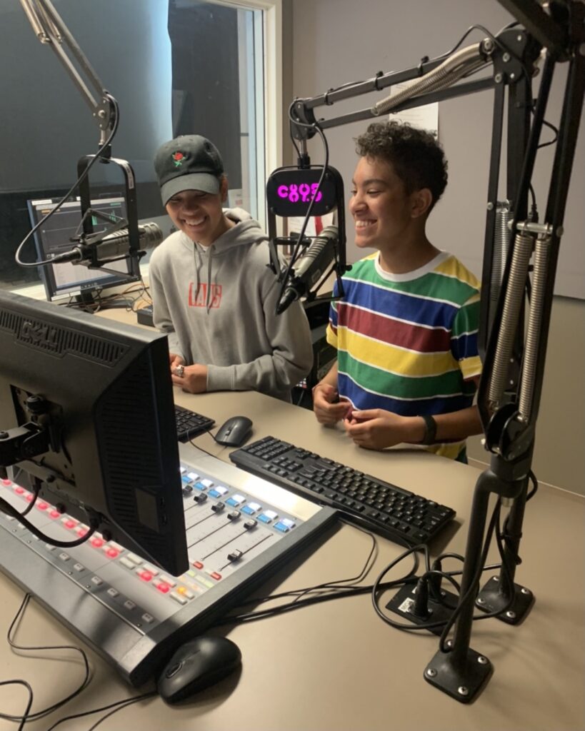 Two people reading from a sheet and speaking into a microphone while wearing headphones in a radio studio.