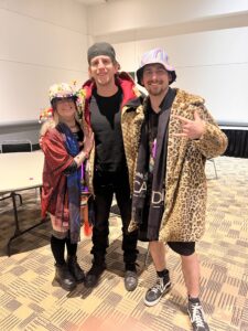 Three people dressed in colorful and eccentric costumes, smiling and posing for a photo at a themed event.