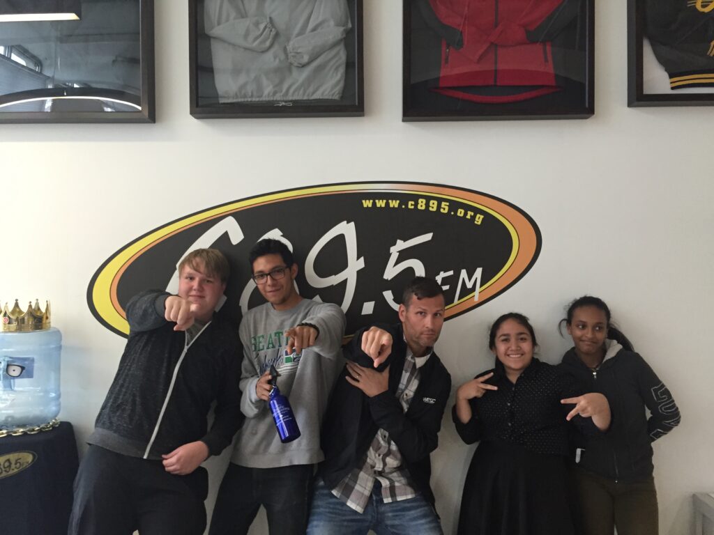 Group of four individuals posing with fun gestures under a C89.5 radio station logo.